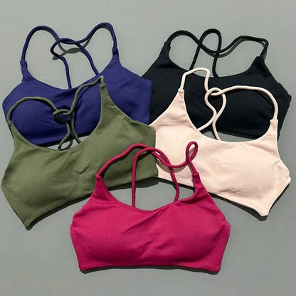 Five sports bras with thin crisscross straps are arranged on a grey surface in purple, olive green, black, light pink, and magenta.