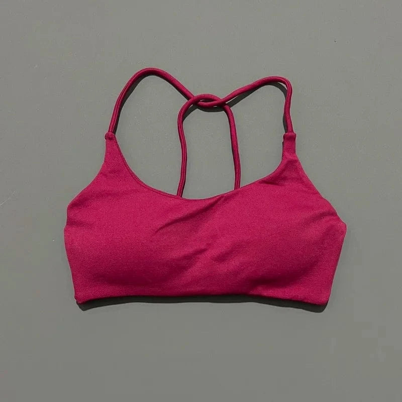 A magenta sports bra with a low scoop neckline and thin, crisscrossed spaghetti straps set against a plain grey background.