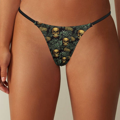 A close-up of thong underwear featuring a dark-toned pattern of skulls nestled within green pineapple foliage and thin black elastic straps.