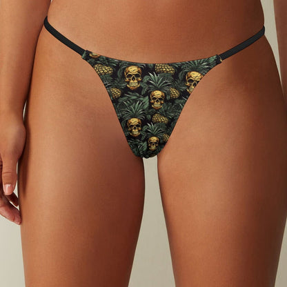 A thong featuring a repeating pattern of gold skulls and green pineapple leaves on a black background, worn by a person.