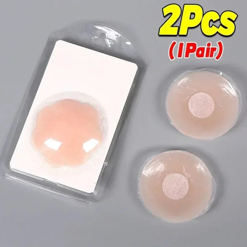 Two circular, beige silicone nipple covers are displayed, with one packaged in a clear plastic case and the other two placed next to it on a grey surface.