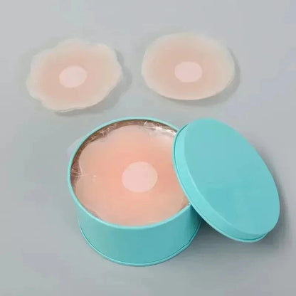 Three silicone nipple covers are displayed, with two placed on a light gray surface and one inside an open, round, light blue metal tin.