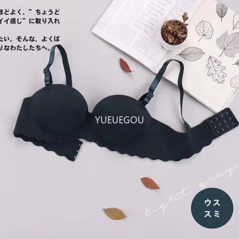 A black strapless bra with scalloped edges is displayed on a flat surface next to an open book, a small potted plant, and three scattered dry leaves.