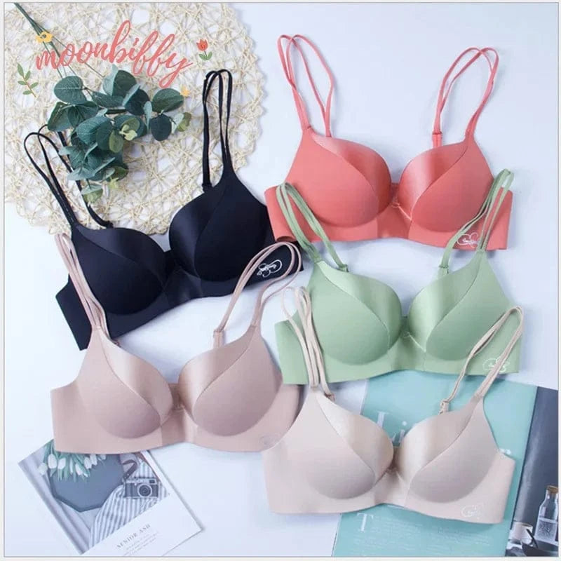 Five seamless bras with thin double straps are arranged on a white surface in black, peach, sage green, and two shades of beige.