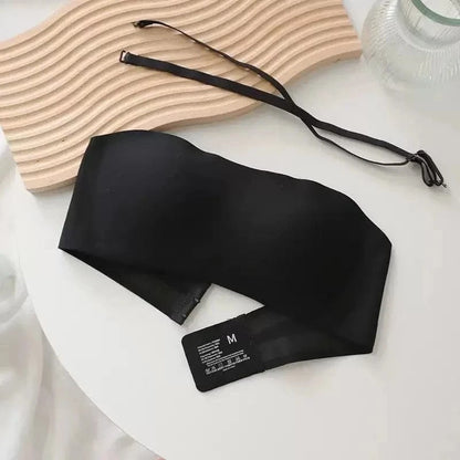 A black strapless bandeau bra with an included detachable shoulder strap laid on a white surface next to a wavy wooden decor piece.
