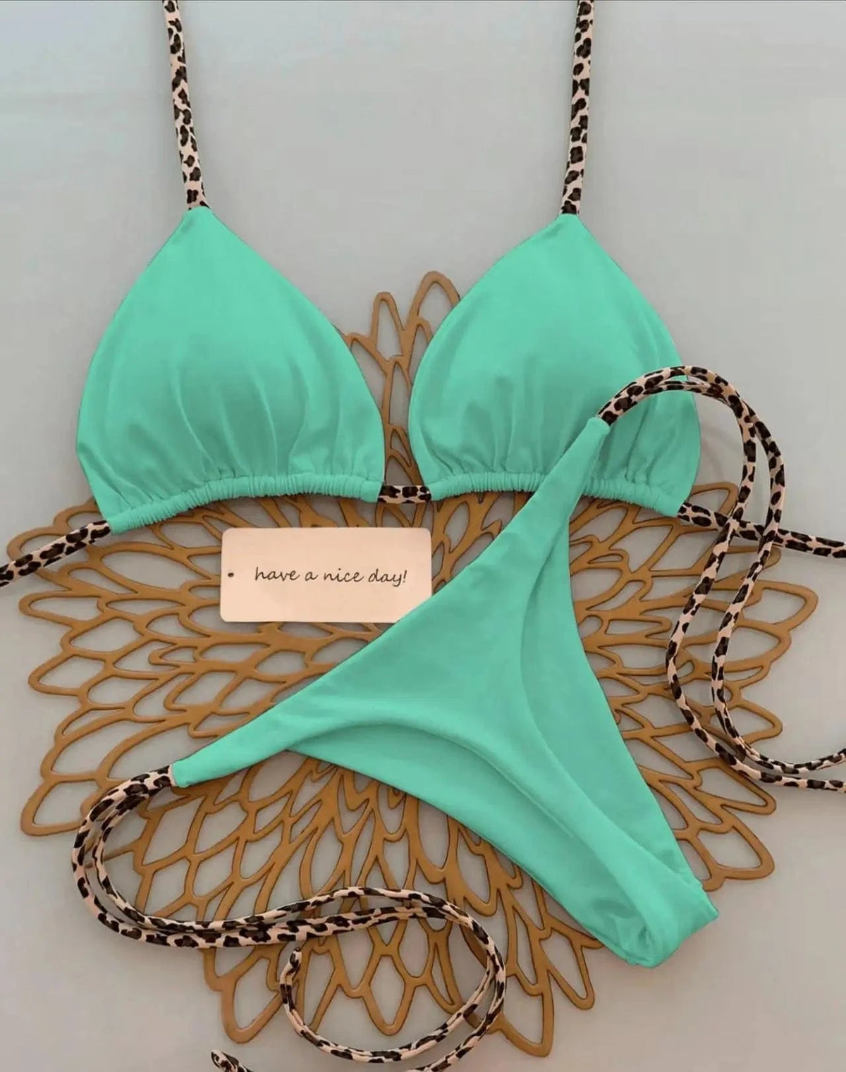 A mint green triangle bikini set featuring leopard print straps, laid out flat on a decorative gold-colored mat with a small card that says have a nice day.