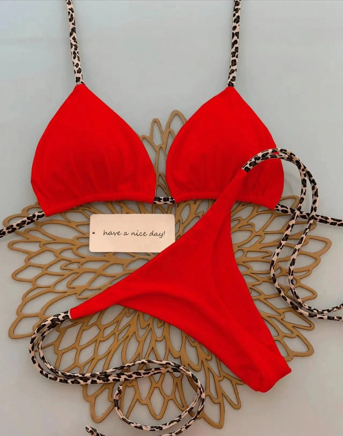 A red bikini set with thin leopard print straps laid out on a gold decorative placemat next to a small note that says have a nice day.