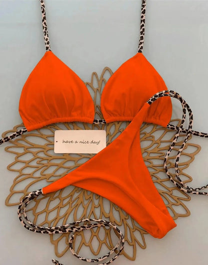 A vibrant orange two-piece bikini featuring thin leopard print straps and ties, laid flat on an intricate gold-toned placemat with a small white note that says have a nice day.