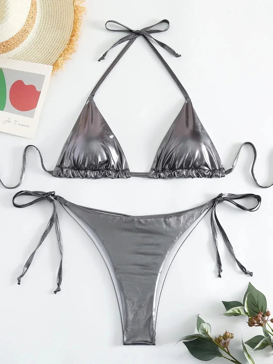 A metallic silver two-piece bikini set consisting of a triangle top with tie-back straps and matching side-tie bikini bottoms, laid flat on a white surface next to a straw hat and green leaves.