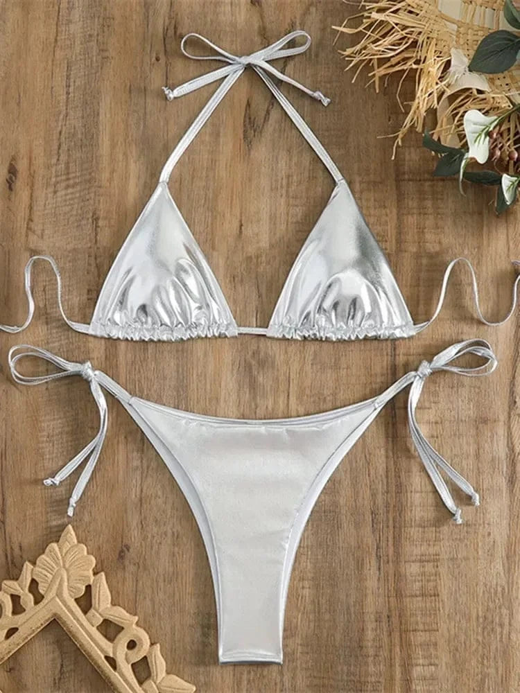 A silver metallic two-piece bikini set laid flat on a wooden surface, featuring a triangle top and tie-side bottoms.