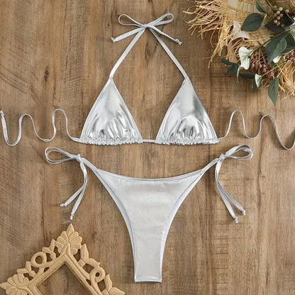 A metallic silver two-piece string bikini set featuring triangular cups and side-tie bottoms laid flat on a wooden surface.