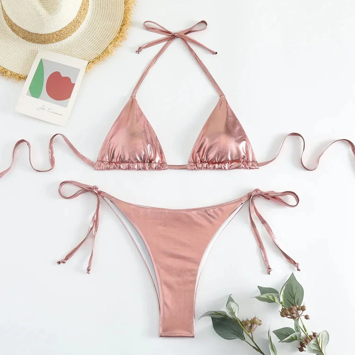 A metallic rose gold bikini set featuring a string triangle top and matching tie-side cheeky bottoms, arranged on a white surface next to a straw hat and a small decorative card.