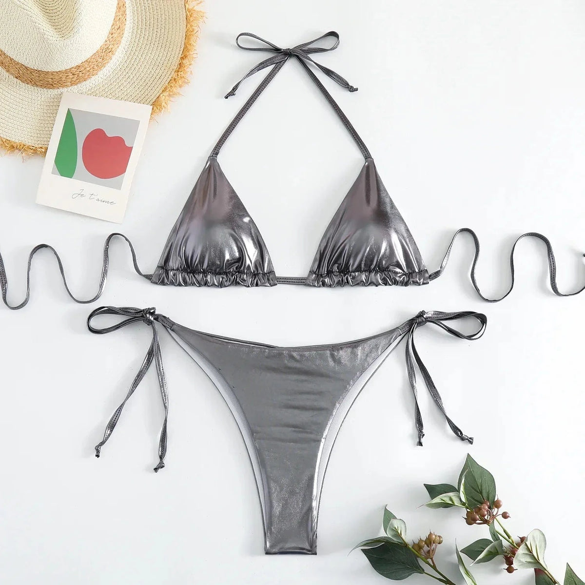 A metallic silver bikini set features a triangle bikini top with tie-back strings and matching side-tie bikini bottoms, laid out on a white surface with a straw hat and a small decorative card.
