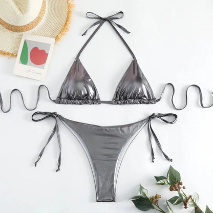 A metallic silver bikini set features a triangle bikini top with tie-back strings and matching side-tie bikini bottoms, laid out on a white surface with a straw hat and a small decorative card.