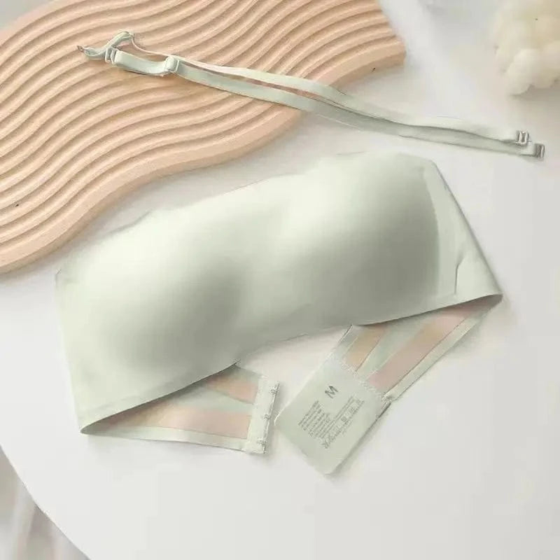 A light sage green strapless bra with a smooth, seamless finish and a set of detachable thin straps lying on a white surface next to a wavy, light-toned textured board.