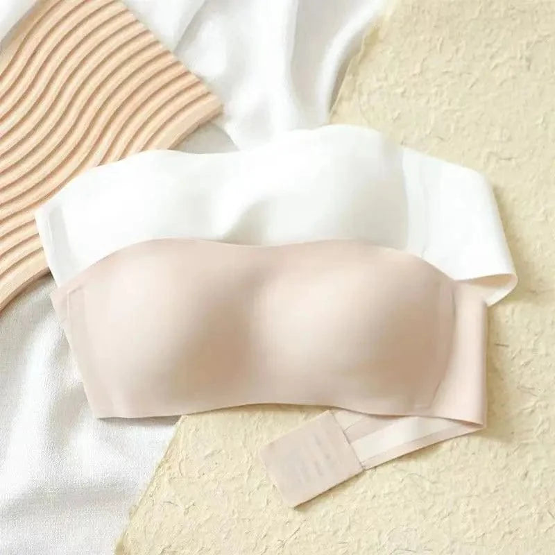 Two strapless bandeau bras in white and beige are layered on a textured surface next to a decorative wooden panel.