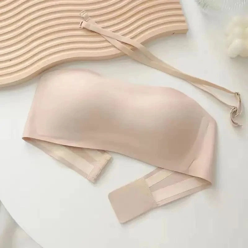 A beige strapless bandeau bra rests on a white surface next to a decorative wavy wooden piece and a separate thin, matching adjustable strap.