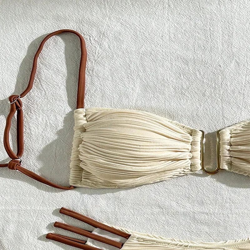 A beige pleated bikini top featuring thin brown adjustable shoulder straps and a gold-toned U-shaped metal connector at the center, laid flat on a white textured surface.