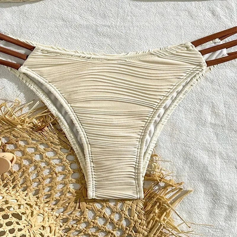A beige textured bikini bottom featuring horizontal ruched fabric and triple brown side straps, laid flat on a woven straw mat.