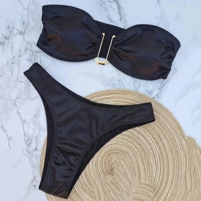 A black two-piece bikini set featuring a strapless bandeau top with a central gold U-shaped metal hardware detail and matching high-cut bikini bottoms, displayed on a marble surface next to a woven straw hat.