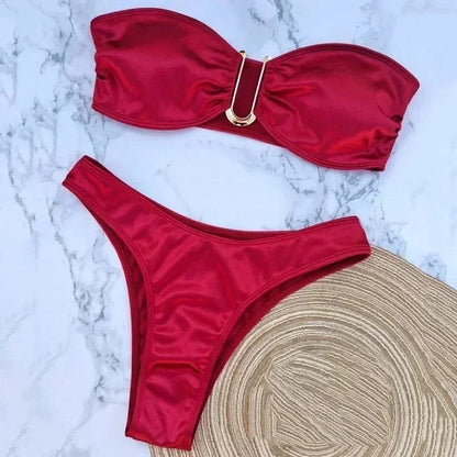 A strapless red bikini set features a U-shaped gold hardware accent in the center of the top and high-cut bottoms, displayed on a marble surface next to a straw hat.
