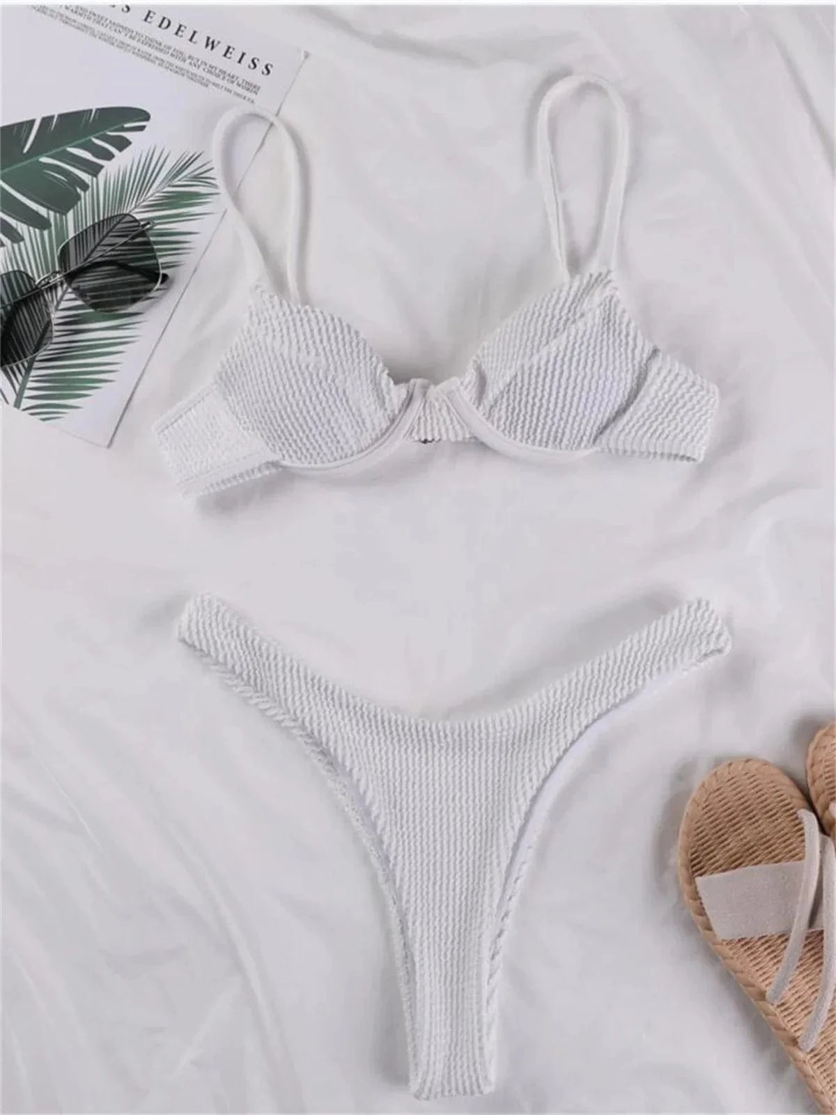 A white textured bikini set with an underwire top and high-leg bottoms laid out on a white surface next to sunglasses and sandals.