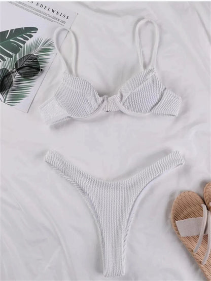 A white textured bikini set with an underwire top and high-leg bottoms laid out on a white surface next to sunglasses and sandals.