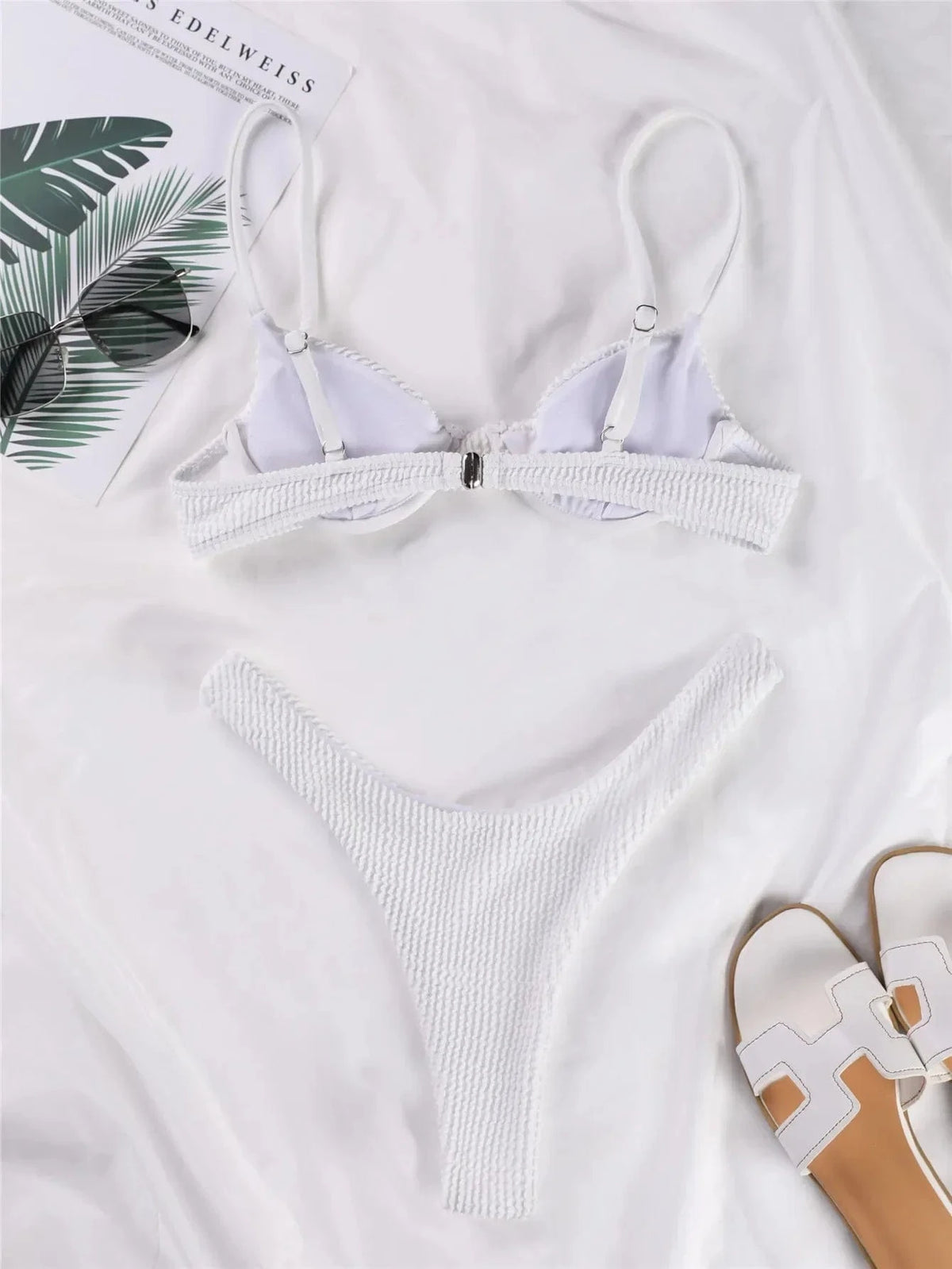 A white textured bikini set with an underwire top and high-cut bottom lies on a white surface next to a pair of sunglasses and sandals.