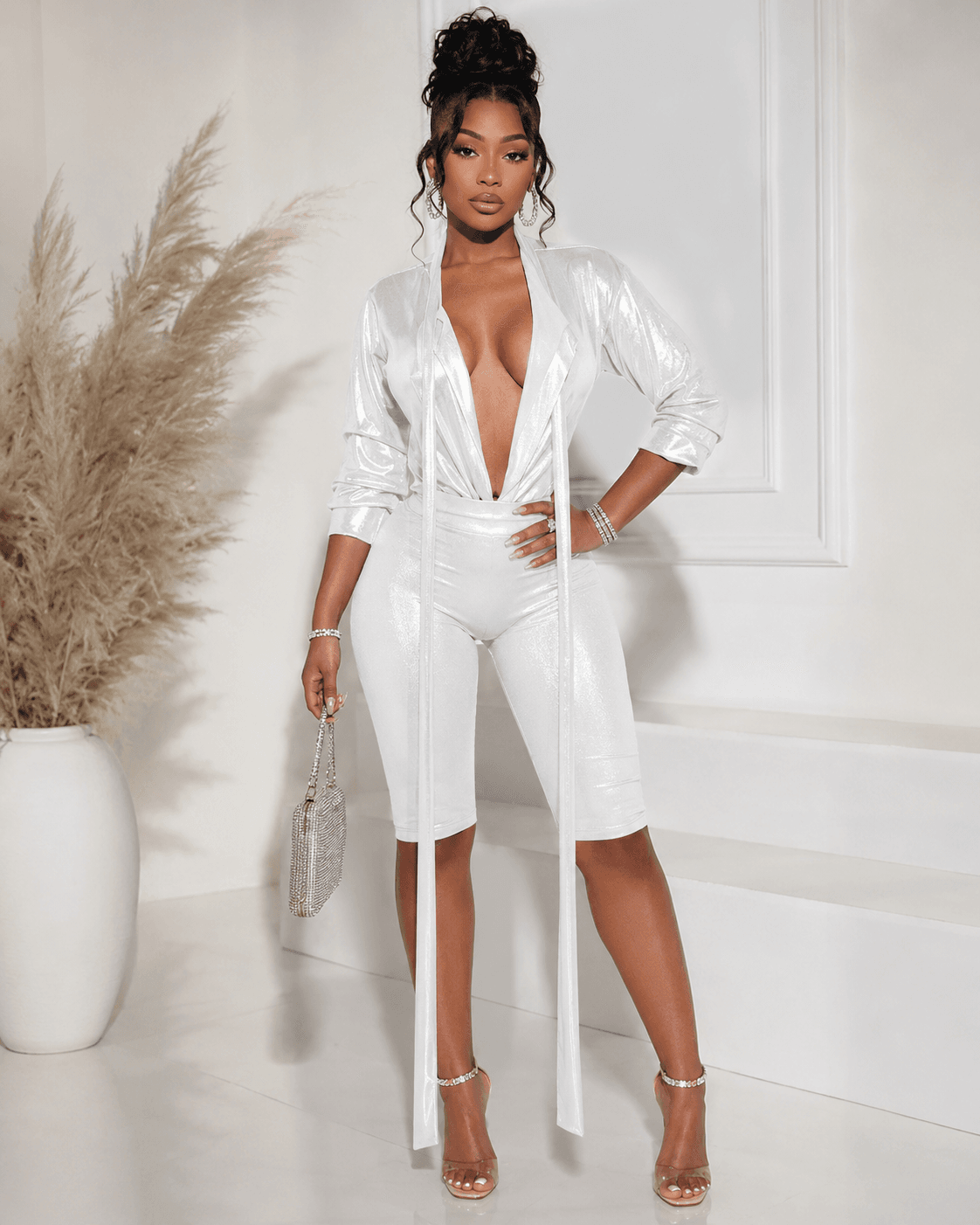 A woman stands posing wearing a metallic silver long-sleeve bodysuit with a deep V-neckline and matching knee-length shorts, paired with clear stiletto heels and a small matching handbag.