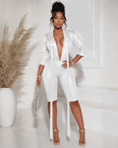 A woman stands posing wearing a metallic silver long-sleeve bodysuit with a deep V-neckline and matching knee-length shorts, paired with clear stiletto heels and a small matching handbag.