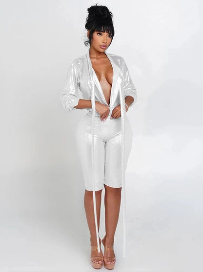 A model poses wearing a shiny white two-piece set featuring a deep V-neck blazer with long hanging strings and matching form-fitting knee-length shorts.
