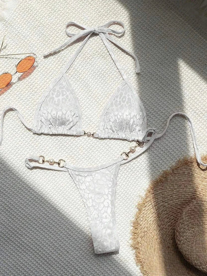 A white animal-print bikini set featuring gold-toned circular hardware with white faux pearls on the straps and center, displayed on a textured white surface next to orange-tinted sunglasses and a straw hat.