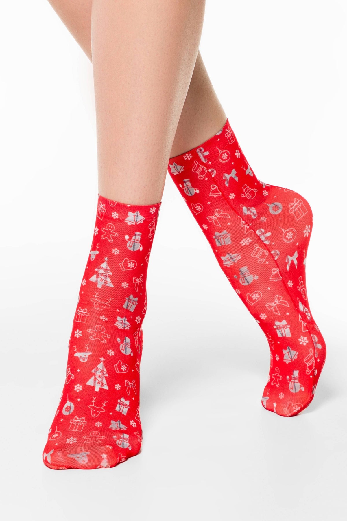Red sheer crew socks featuring a festive pattern of white Christmas trees, snowmen, gingerbread men, gift boxes, bows, and snowflakes worn on a person's legs against a white background.