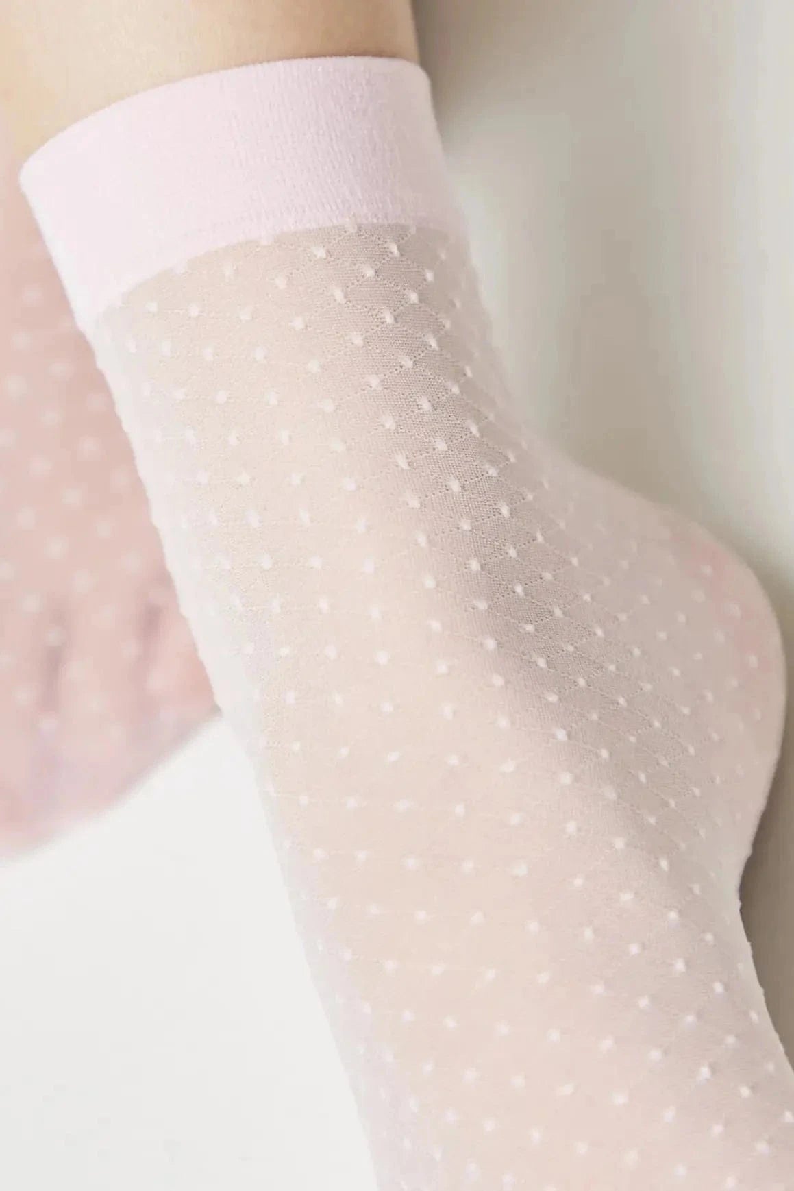 A close-up of a foot wearing a sheer, light pink sock featuring a delicate white swiss dot pattern and a solid pink cuff.