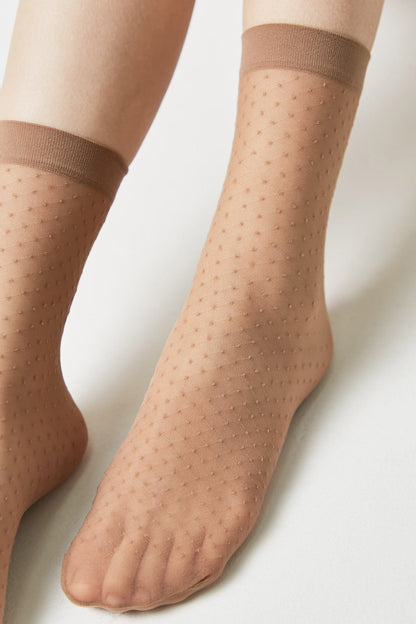 A pair of sheer tan ankle socks featuring a subtle Swiss dot pattern and a darker brown ribbed cuff, worn on a person's feet against a plain white background.