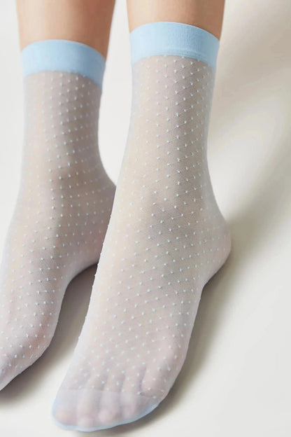 A pair of sheer white ankle socks featuring a subtle Swiss dot pattern and light blue trim at the cuff, worn on a person's feet against a plain white background.