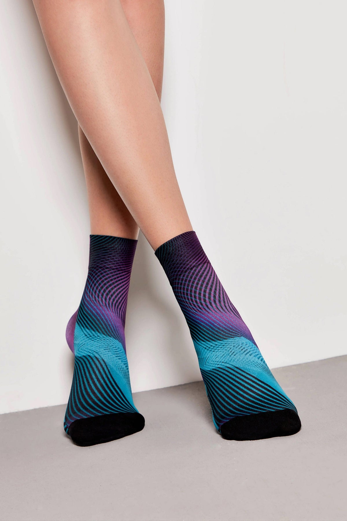 A pair of crew-length socks featuring a vibrant abstract pattern of wavy purple, blue, and black lines with solid black toe caps, worn by a model against a neutral background.