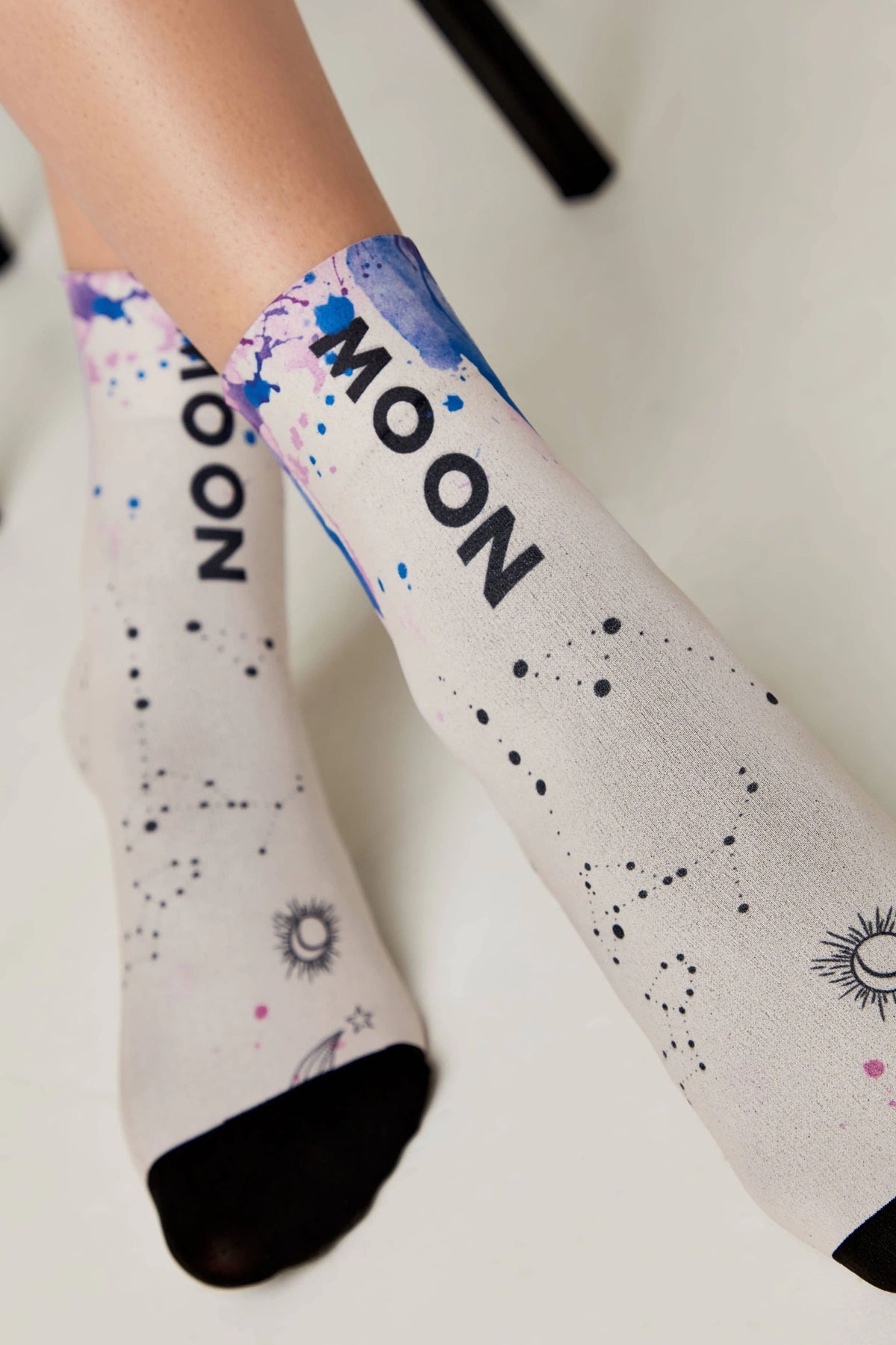 A pair of crew socks featuring a light grey base, watercolor-style blue and purple accents, the word MOON printed in black, and small black celestial illustrations of constellations, stars, and suns.