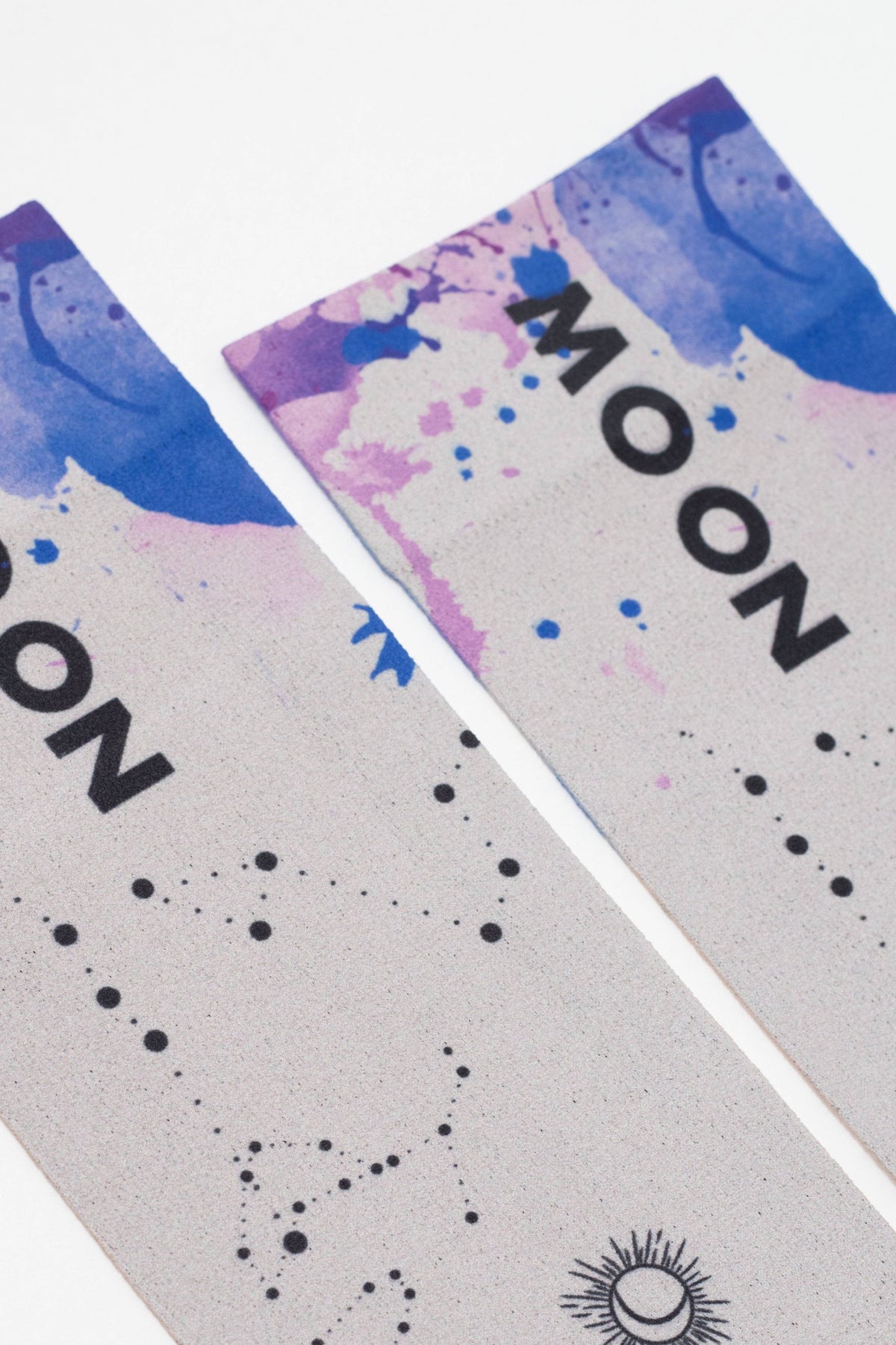 Two rectangular bookmarks featuring a watercolor design of blue and purple splashes above a grey base with black constellation patterns and the word MOON printed in bold black text.