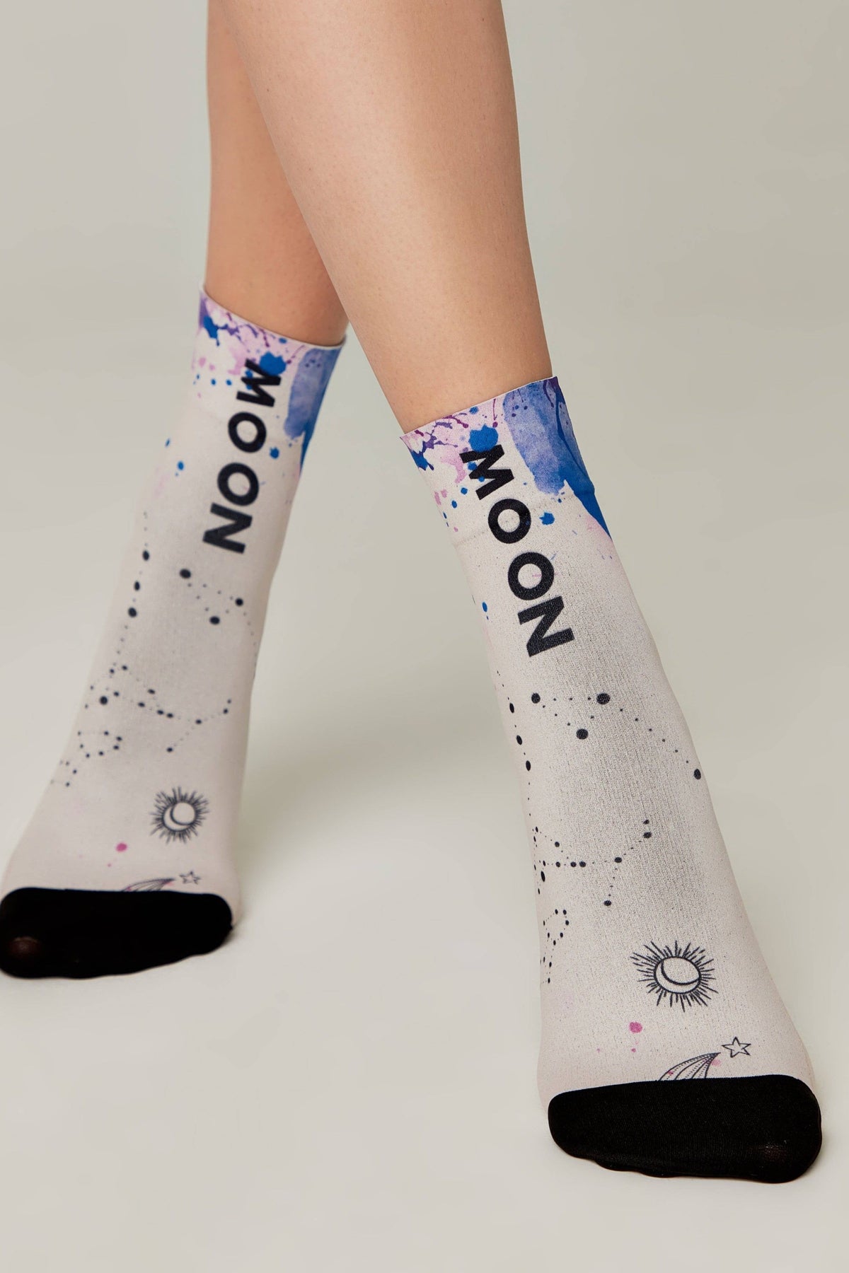 A pair of light-colored socks featuring a blue watercolor design, black star and sun motifs, and the word MOON printed vertically on each side with contrasting black toe sections.
