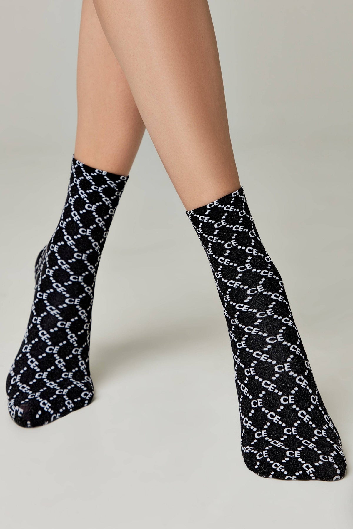A pair of black ankle socks with a repeating white monogram pattern featuring the letters CE against a subtle shimmering background worn on a person's feet.