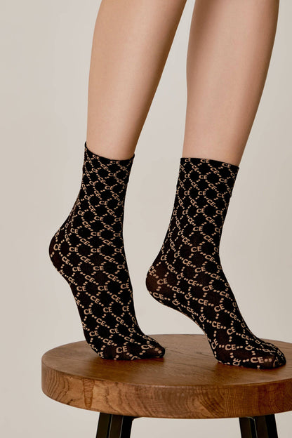 Sheer black socks with a repeating tan diamond grid pattern containing the letters CE are worn on a person's feet as they balance on a small wooden stool against a neutral background.