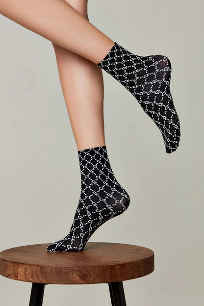 A pair of sheer black crew socks patterned with a white repeating logo are worn on a person's feet, with one foot resting on a round wooden stool and the other lifted in the air against a neutral background.