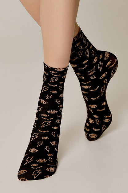 A pair of black socks with a repeating pattern of gold lightning bolts and stylized open eyes worn on a person's feet against a plain background.
