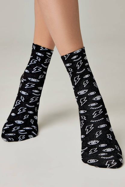 A pair of black crew-length socks features a repeating white pattern of stylized lightning bolts and eyes with long eyelashes worn on a person's feet.