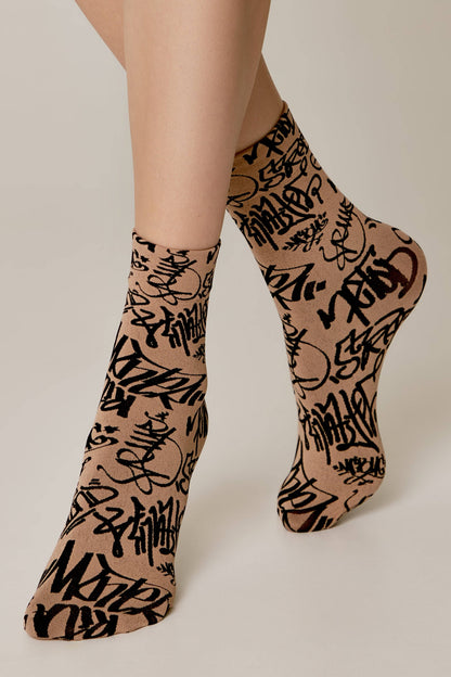 A pair of tan-colored ankle socks with a black graffiti-style text print worn on a person's feet against a light background.