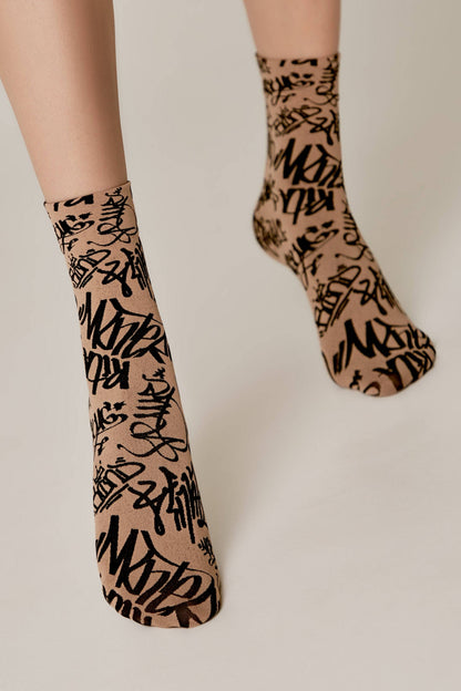 A pair of tan ankle socks featuring an all-over black graffiti-style print being worn on a person's feet.