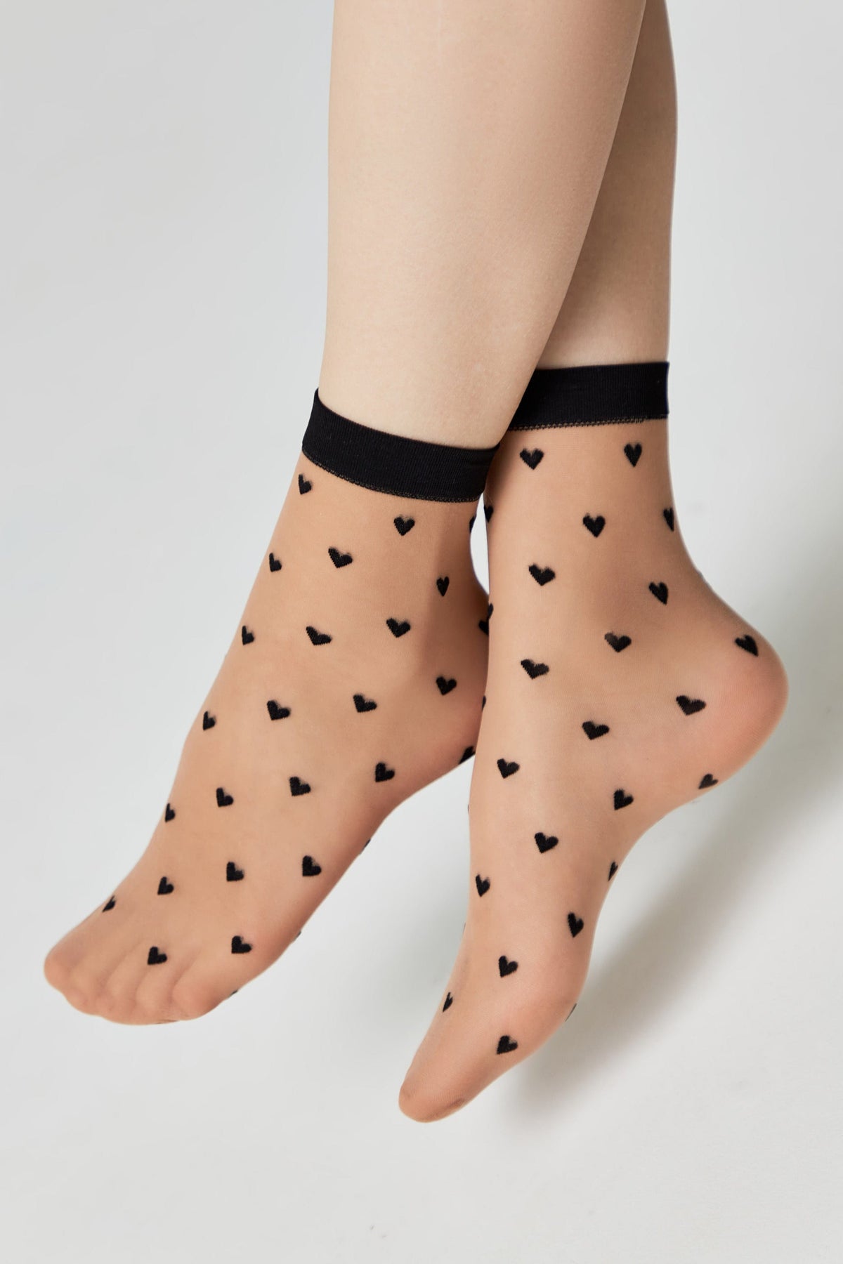 Sheer beige ankle socks patterned with small black hearts, featuring a solid black trim at the top and worn on a pair of legs.