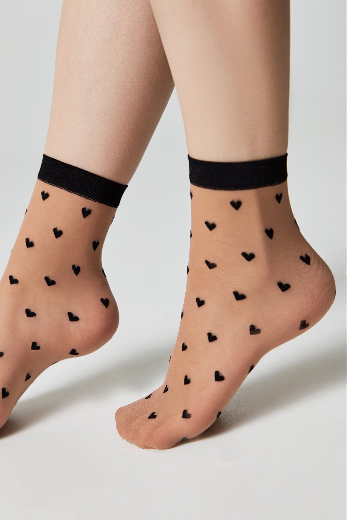 Sheer ankle-high socks featuring a pattern of small black hearts against a beige background with a contrasting black ribbed cuff.