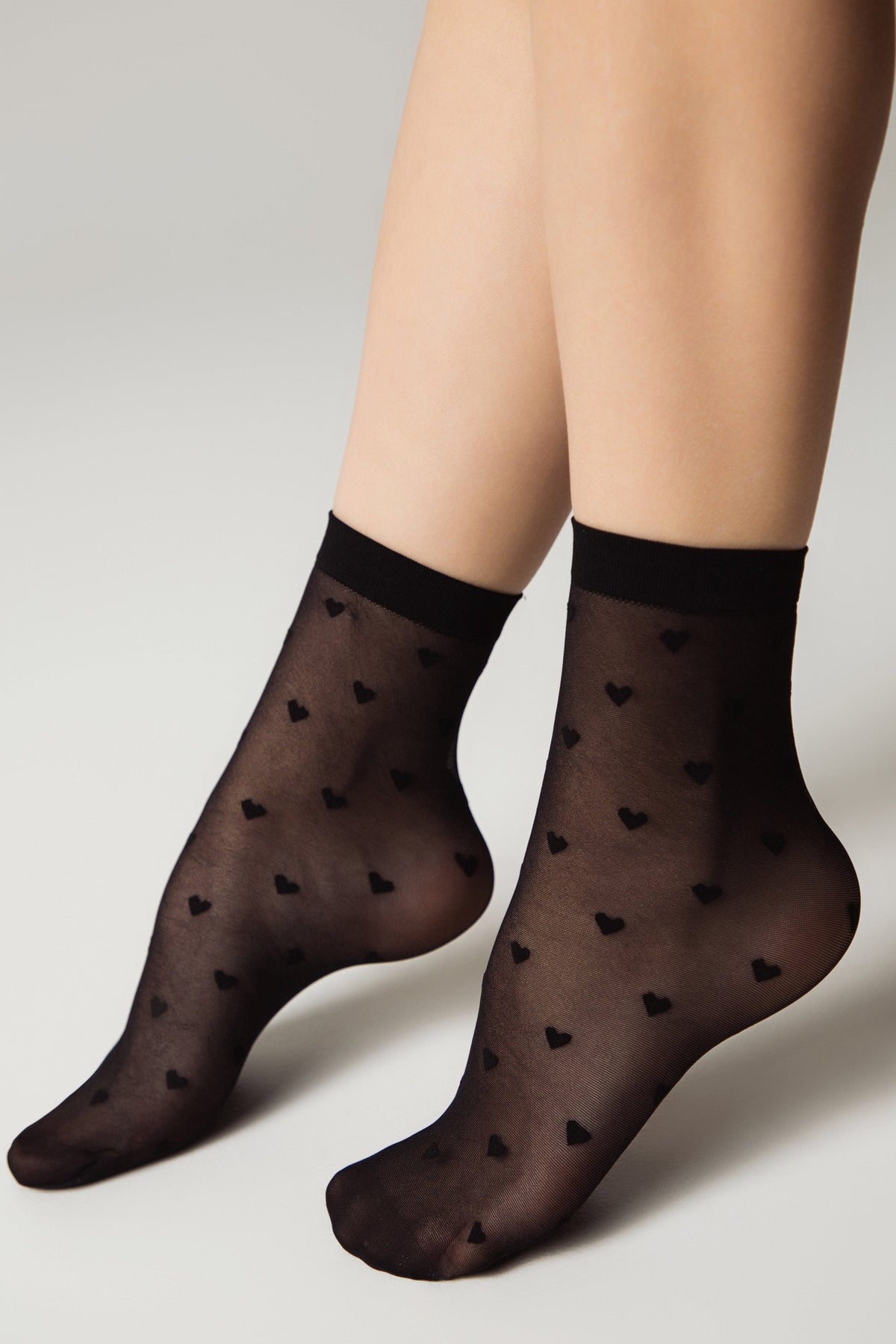 Sheer black ankle socks patterned with small black hearts worn on a pair of legs against a plain light background.
